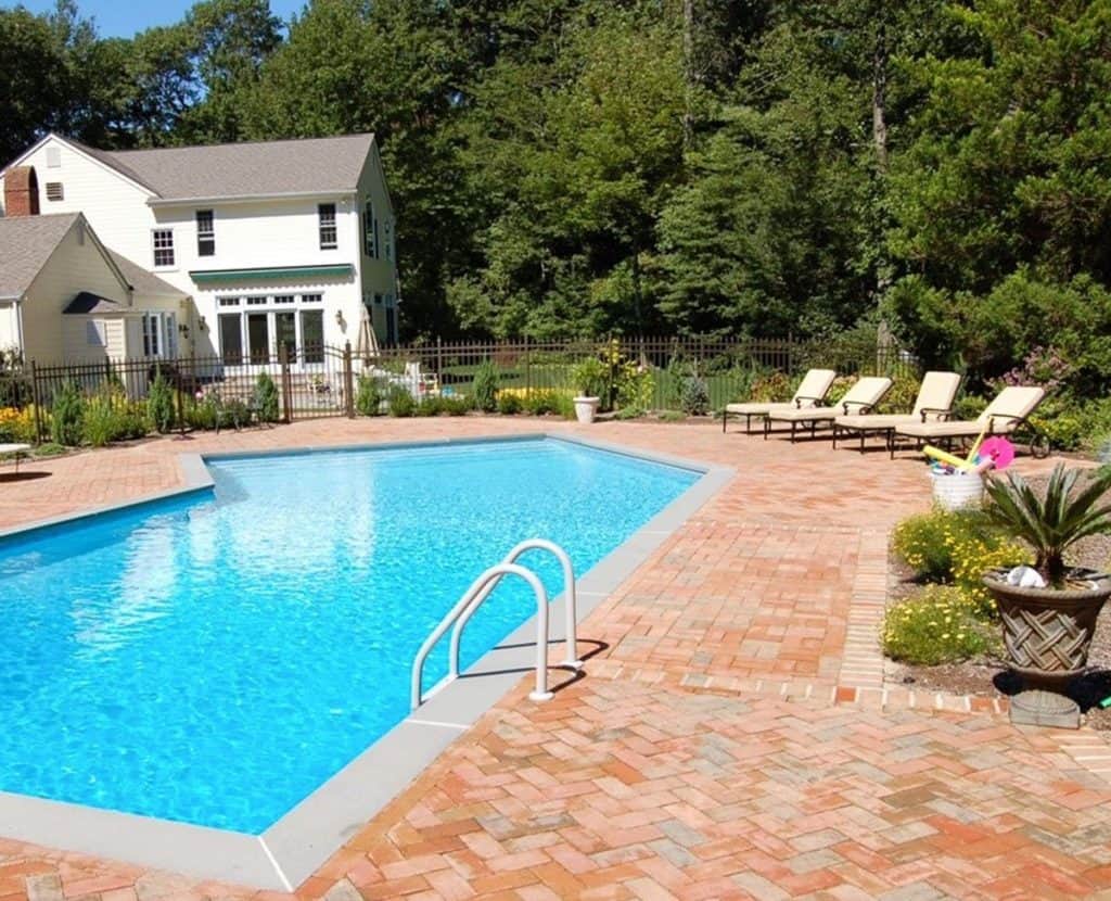 A backyard swimming pool with clear blue water is surrounded by a brick patio. Four lounge chairs are set up on the right side. A yellow house with a white fence and trees is in the background, showcasing meticulous care that reflects expert pool services in Suffolk County, NY.
