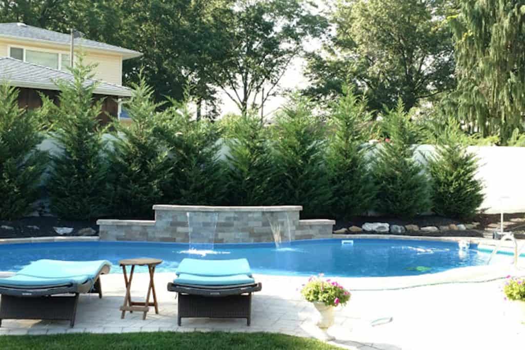 A sunny backyard boasts a round swimming pool, ideal for those considering pool installation in Nassau County, NY. Two loungers with blue cushions and a small wooden table adorn the patio. A beautiful water feature enhances the pool's charm, while tall trees and bright flowers frame this serene oasis.