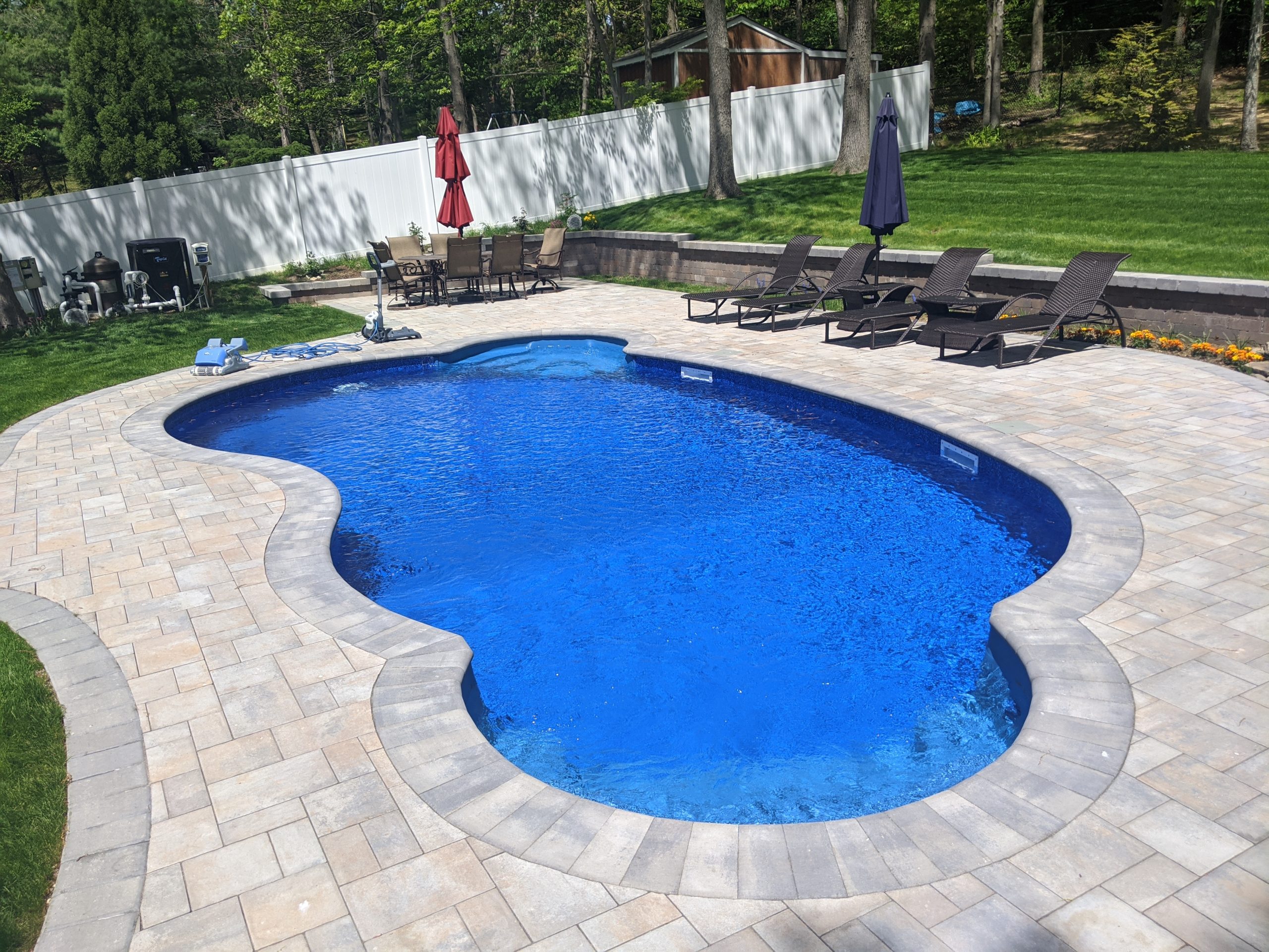 A kidney-shaped inground swimming pool with clear blue water is surrounded by a stone patio. Lounge chairs with umbrellas and a grassy lawn are in the background, bordered by a white fence and trees.