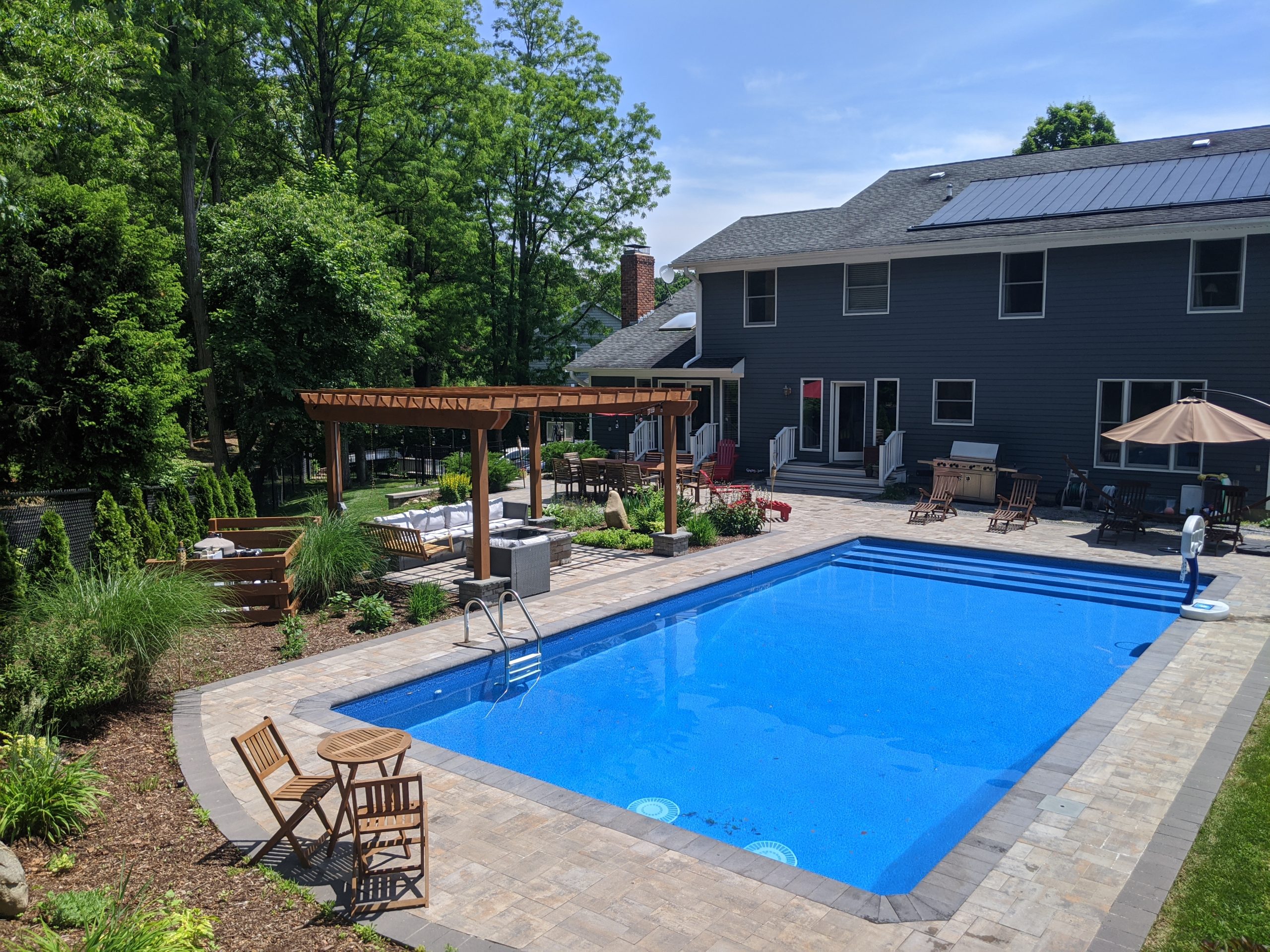 A spacious backyard features a large rectangular swimming pool, lounge chairs, a wooden pergola, outdoor seating areas, and lush landscaping next to a two-story house on a sunny day.
