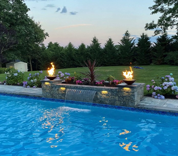A backyard pool with clear blue water, a stone water feature, two fire bowls, and colorful flowers. Tall trees and a grassy lawn are in the background at sunset.