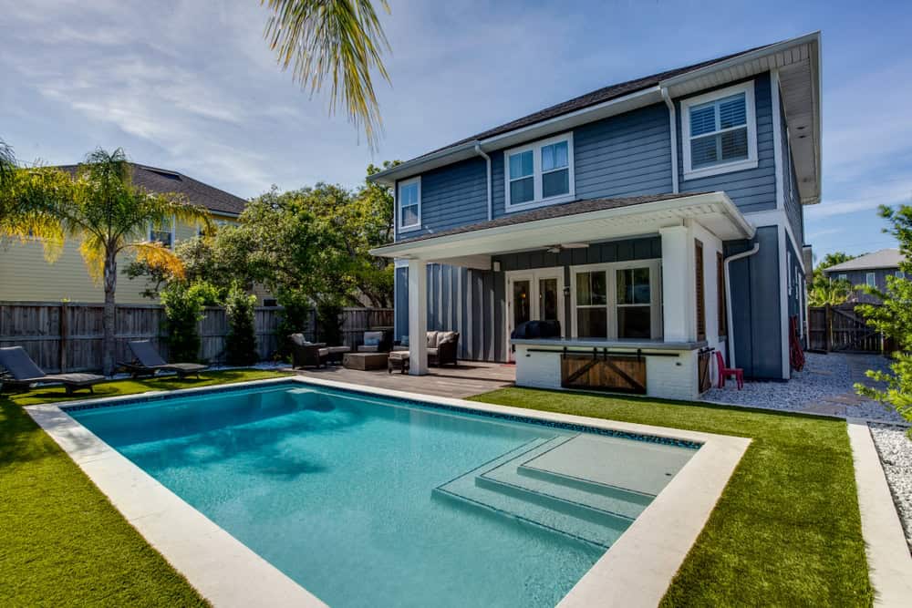 Two-story house with gray siding, a covered patio, and a backyard pool installed by professionals in Nassau County, NY. Surrounded by green grass, palm trees, and a wooden fence, the outdoor area includes lounge chairs and a grill on the patio. The sky is clear and sunny.