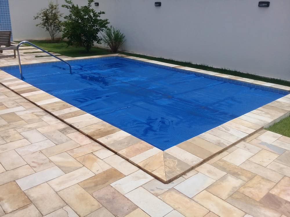 A small, rectangular swimming pool with a blue cover is surrounded by a light-colored stone patio. Green plants and grass create a serene backdrop, while a metal handrail extends into the pool—ideal for those considering pool installation in Nassau County, NY.