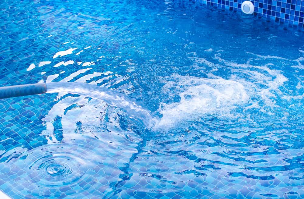 Water flows into a swimming pool from a hose, creating ripples and bubbles on the surface. The pool's blue tiles reflect the sunlight, enhancing the vibrant blue color of the water. For top-notch maintenance, consider pool services in Suffolk County, NY.
