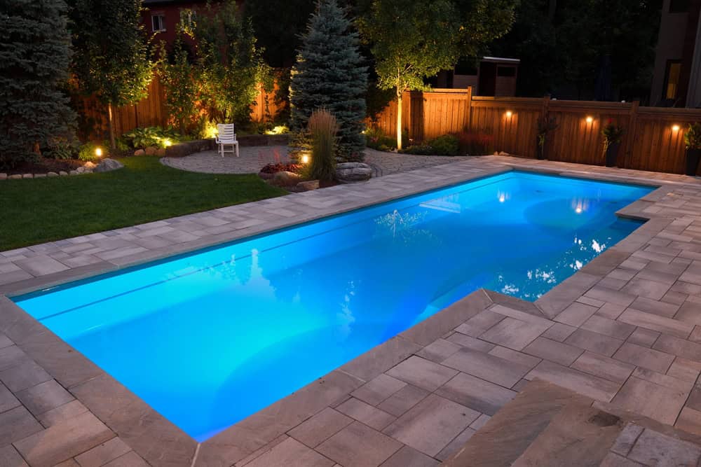 A rectangular swimming pool glows with blue underwater lights at night. Surrounded by a paved deck and lush landscaping, including trees, shrubs, and a wooden fence, it epitomizes elegance. Enjoy peaceful nights or enhance your space with expert pool installation in Nassau County NY.