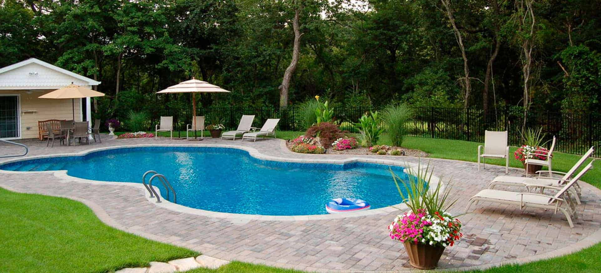 A backyard oasis features a gracefully curvy swimming pool built to the standards of top-notch pool installations in Suffolk & Nassau County, NY. Surrounded by a stone patio, cozy lounge chairs, and umbrellas, the lush greenery and flowering plants create a serene and natural setting.