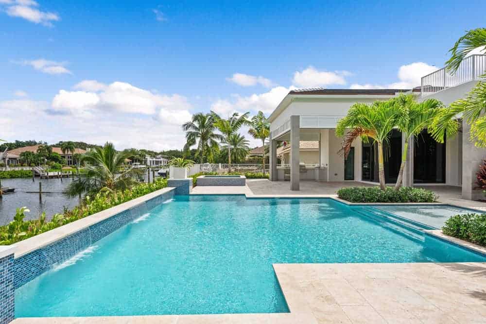 Luxury home with a large, blue swimming pool overlooking a scenic waterfront. Palm trees and modern architecture enhance the view. Expert pool installations in Suffolk & Nassau County, NY ensure the design blends seamlessly with the serene and tropical atmosphere.
