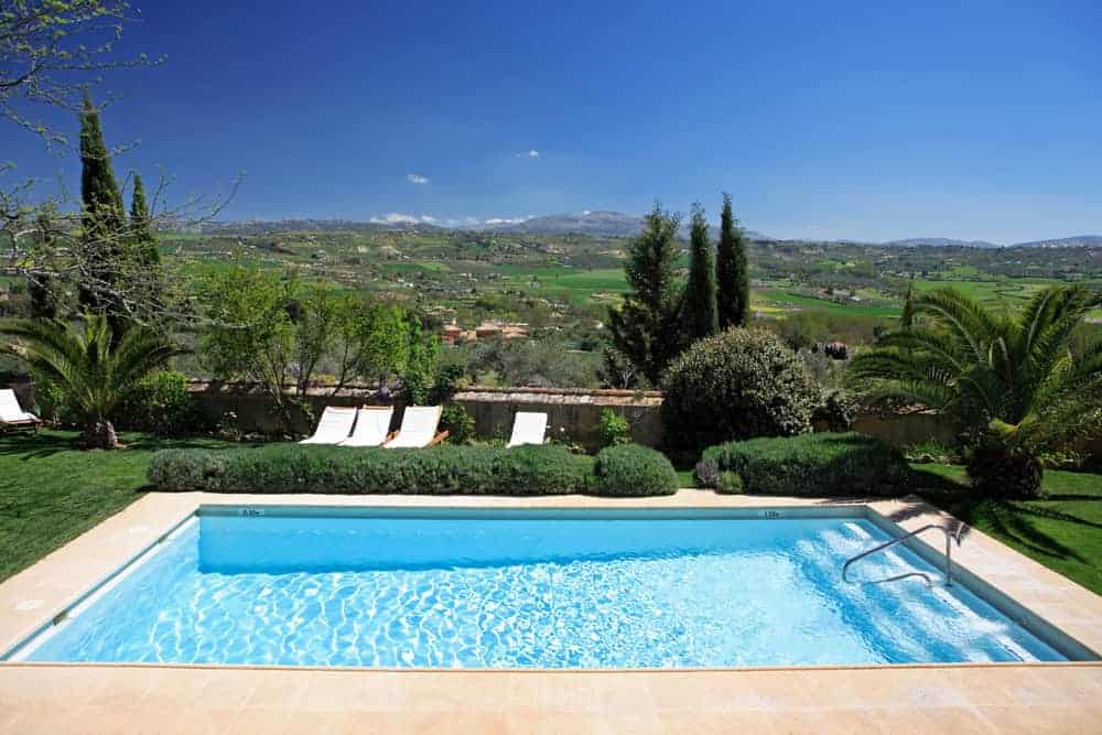 A rectangular swimming pool with clear blue water sits in a landscaped garden with lounge chairs, palm trees, and lush greenery, overlooking a scenic valley and distant hills under a bright blue sky.