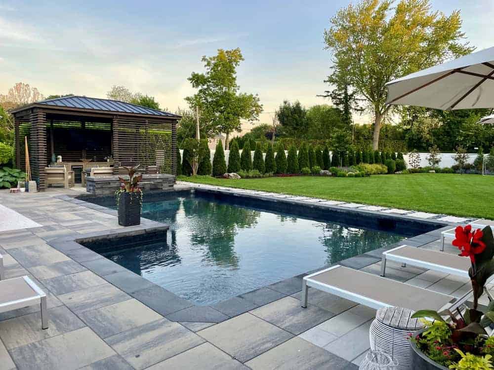 Modern backyard with a rectangular swimming pool, lounge chairs, a shaded pergola, green lawn, potted plants, and tall trees and shrubs providing privacy along the fence.