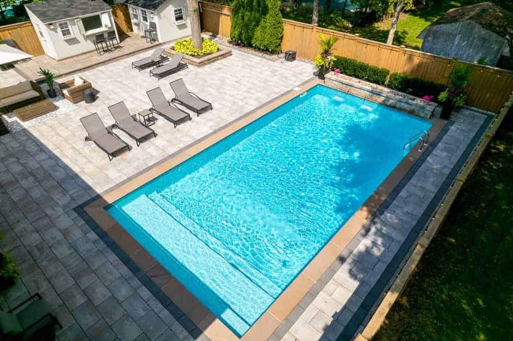 Aerial view of a backyard with a rectangular swimming pool, surrounded by lounge chairs, planters, a patio area, and a wooden fence. There are two small sheds and green landscaping around the pool.