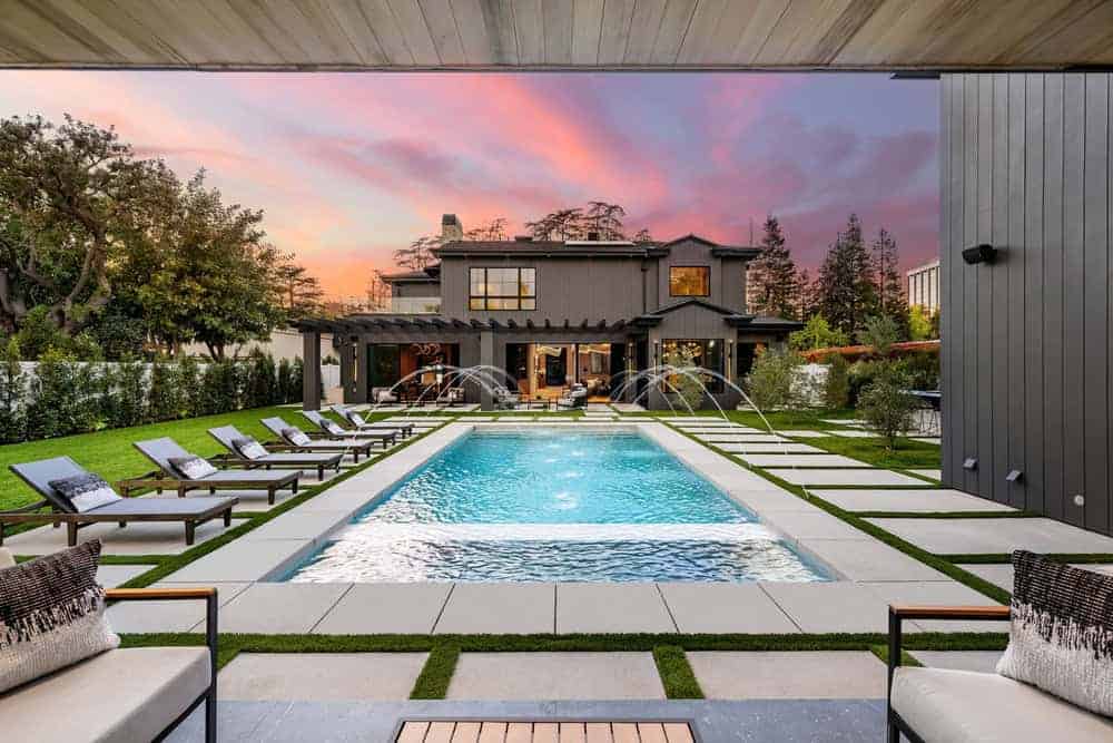 Modern backyard with a rectangular pool, lounge chairs, and water fountains, set against a two-story house at sunset with a vibrant sky. Neatly manicured lawn and contemporary outdoor furniture surround the pool.
