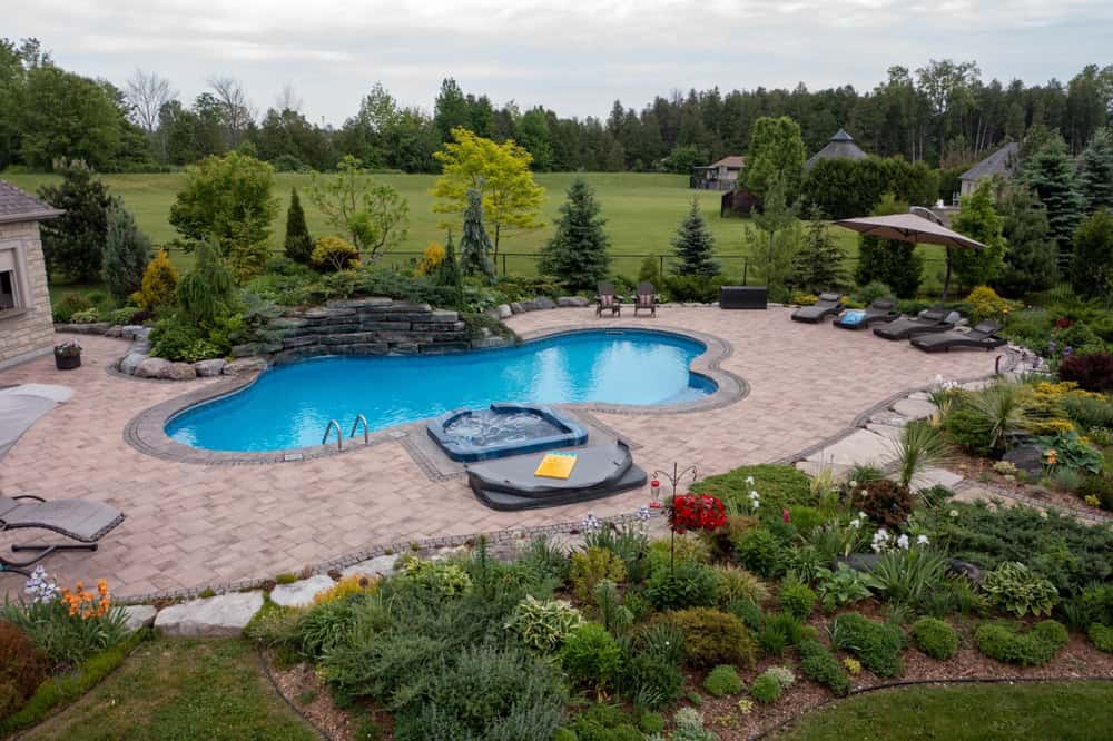 A beautifully landscaped backyard features a curvy swimming pool with a connected hot tub, surrounded by a stone patio, lounge chairs, umbrellas, gardens, and a large grassy field bordered by trees.