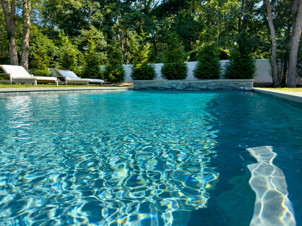 A sparkling blue swimming pool with clear water, built by a top pool contractor in Suffolk and Nassau County, NY. Surrounded by green trees and shrubs, three white lounge chairs rest on the shady deck in the background.
