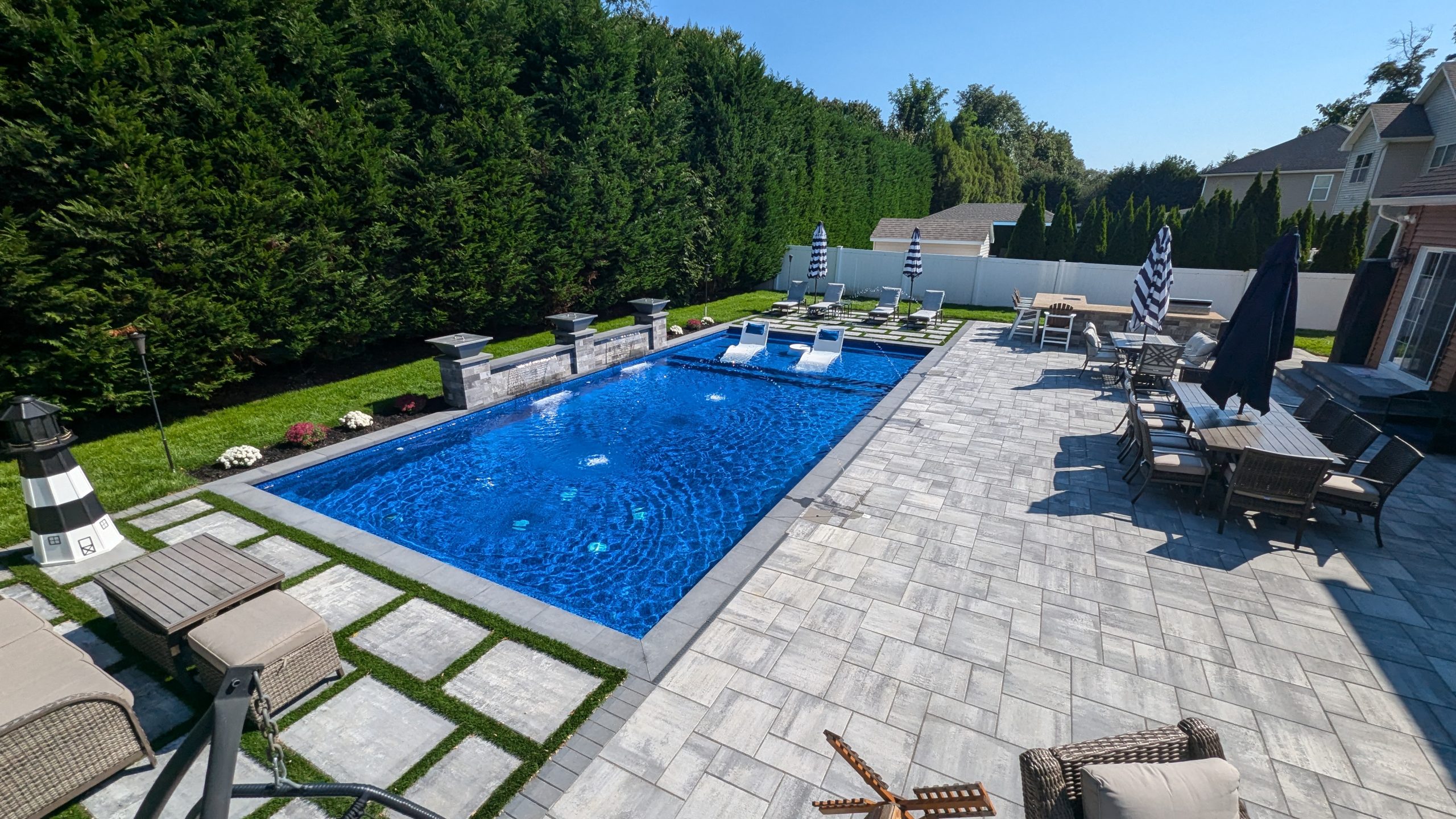 A rectangular backyard swimming pool with lounge chairs in the water, surrounded by stone patio, outdoor dining area, umbrellas, and green hedges under a clear blue sky.